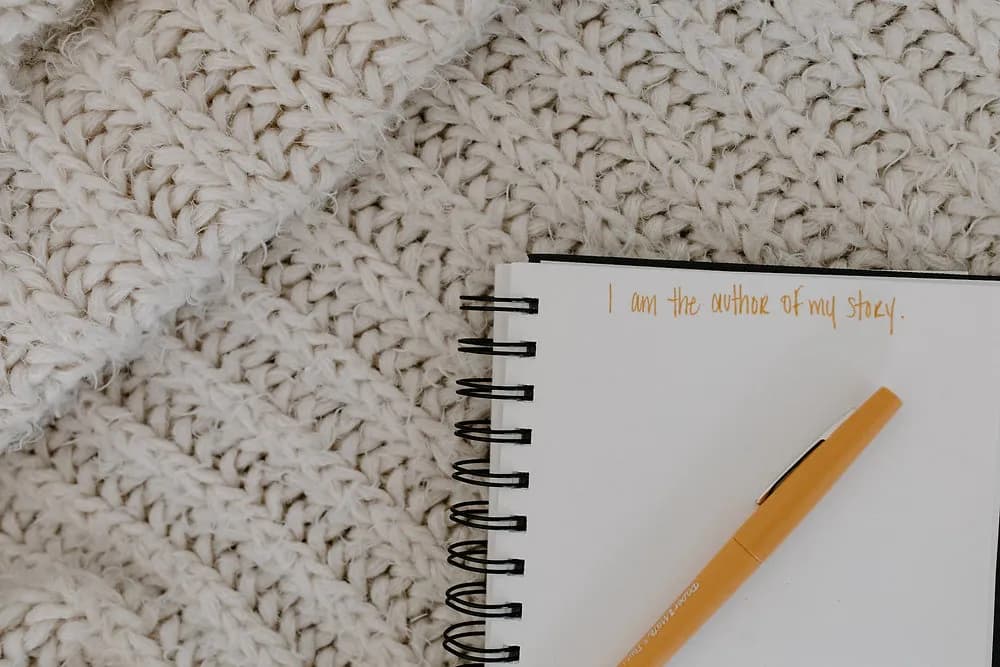 Open spiral notebook on a cream knit blanket with a gold pen and the handwritten line 'I am the author of my story'