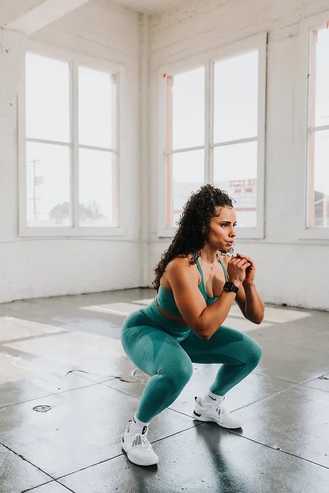 Woman in teal activewear doing a deep squat in a bright, sunlit studio