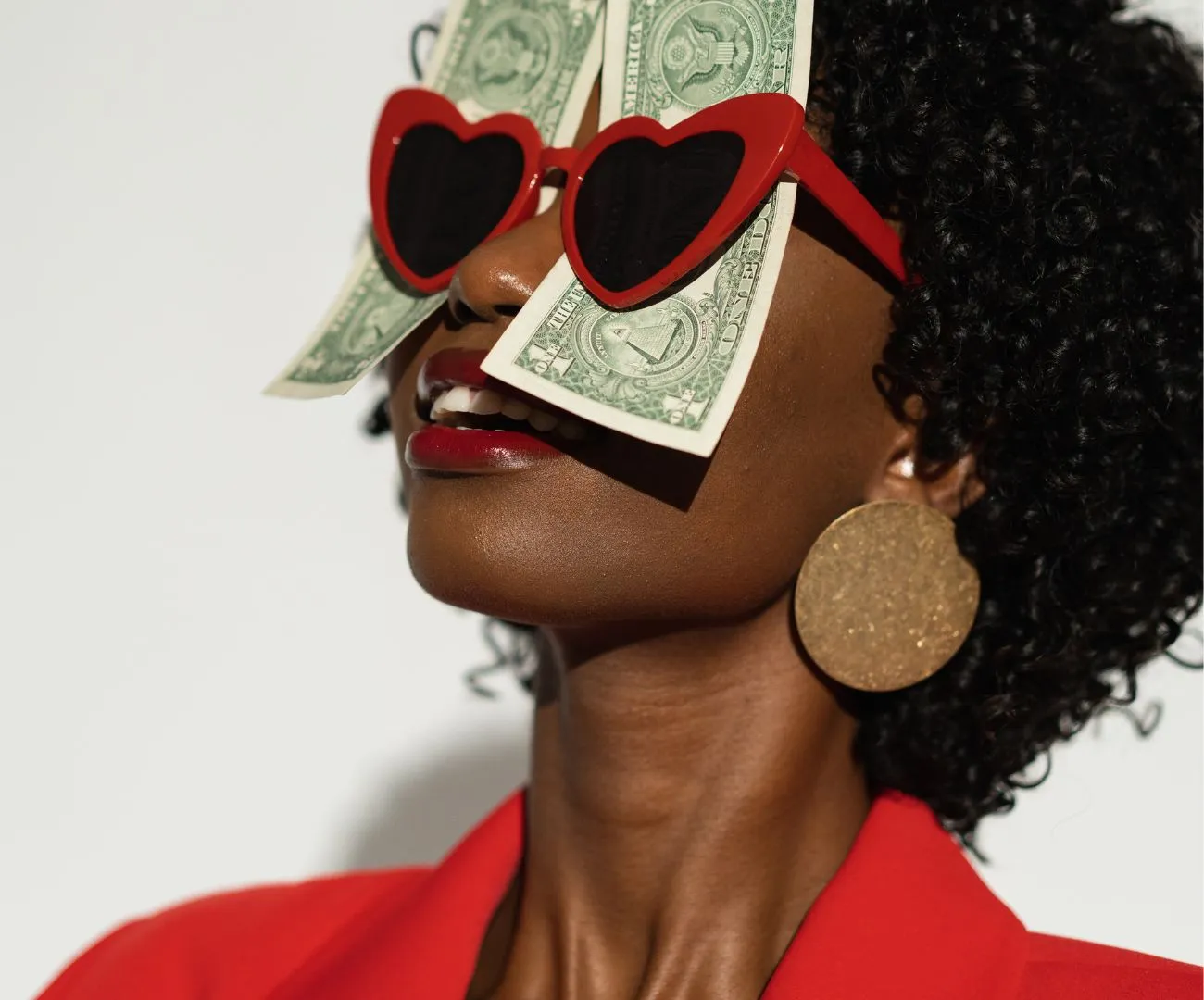 A smiling woman in a red suit wearing heart-shaped sunglasses with dollar bills, symbolising a joyful money mindset