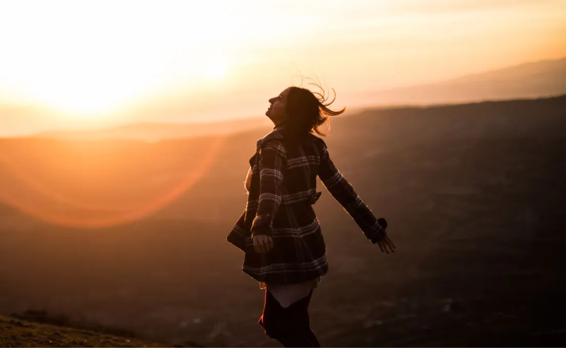 A woman on a hilltop at sunrise, arms open, breathing in the golden light