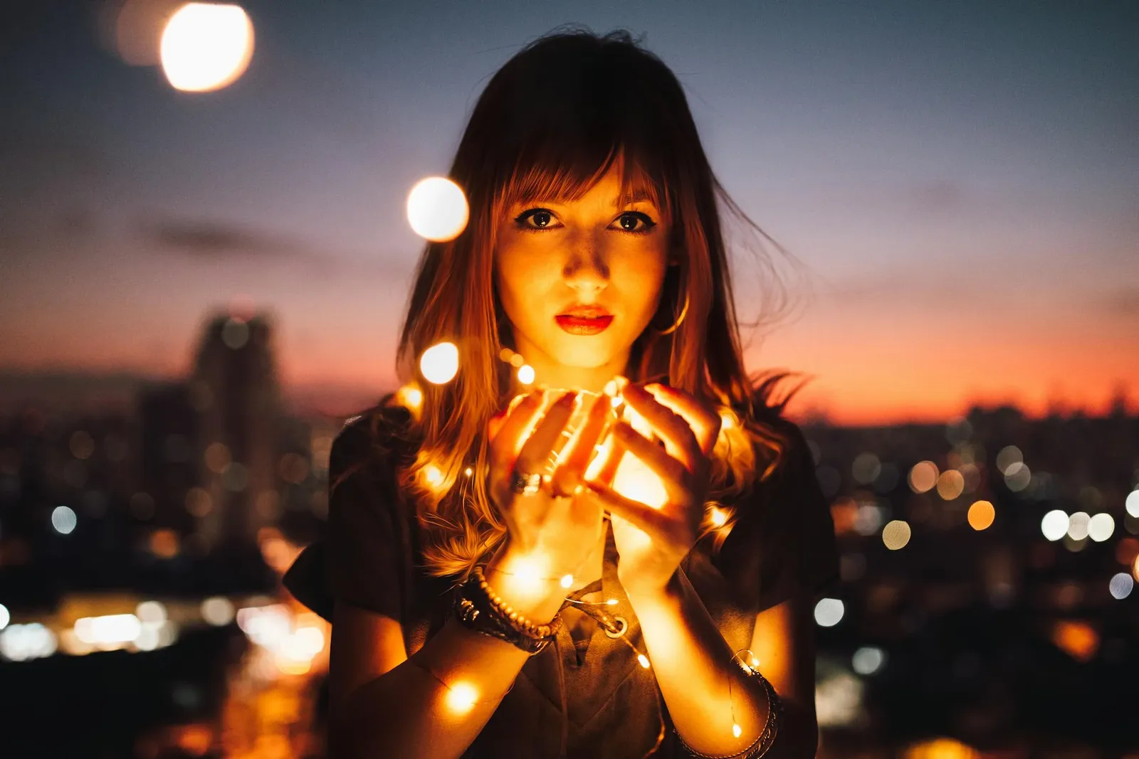 A woman at dusk holding a string of glowing fairy lights above a city skyline