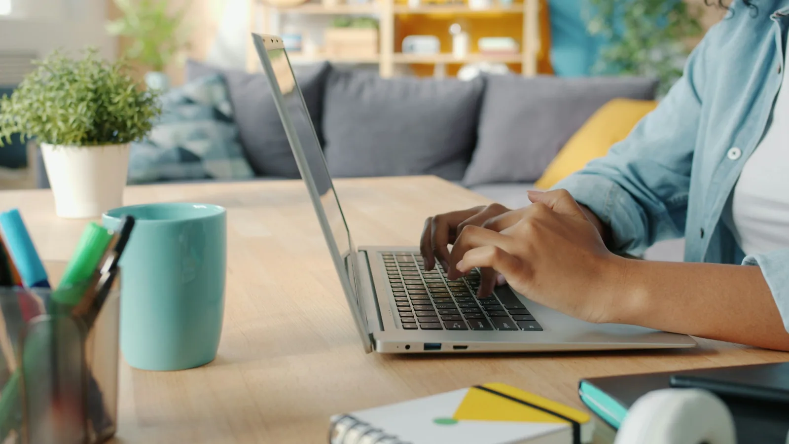 Hände tippen auf einem Laptop am Schreibtisch, daneben eine Pflanze und ein Becher mit Stiften