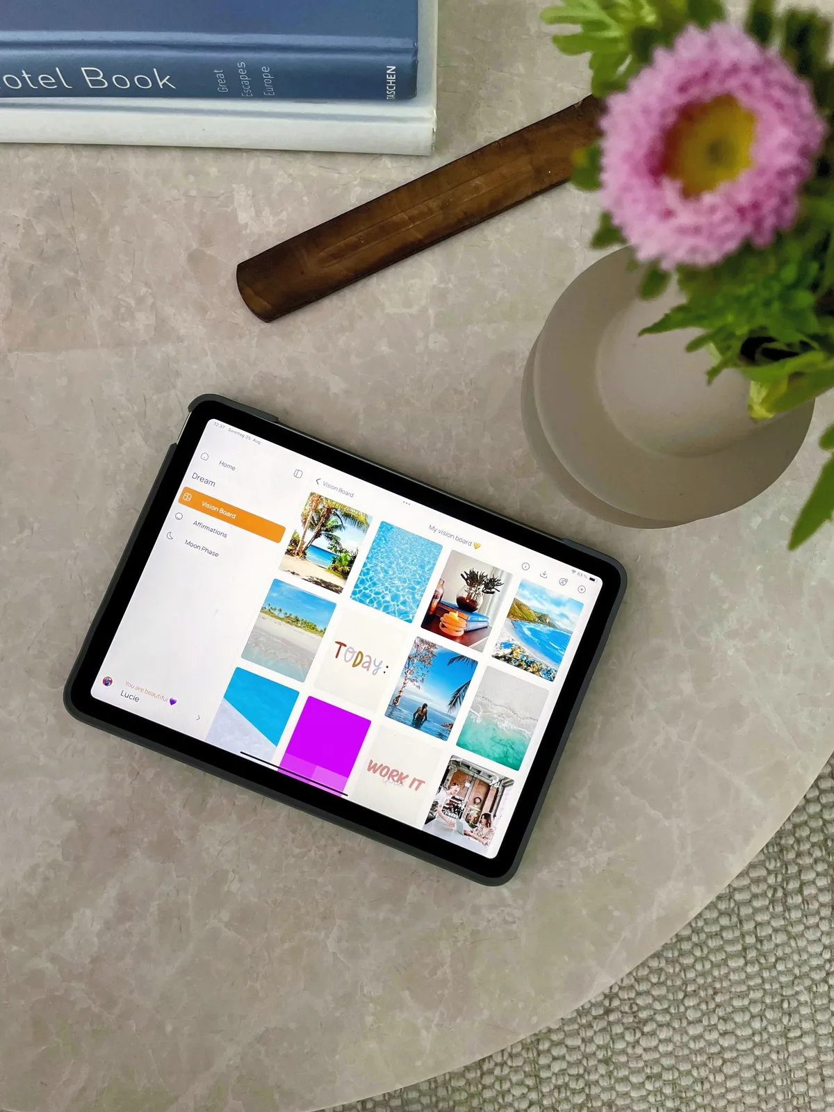Ein Tablet zeigt ein farbenfrohes digitales Vision Board mit Strand- und Reisefotos in der VISIYA App, auf einem Steintisch neben einem Buch und einer Blumenvase
