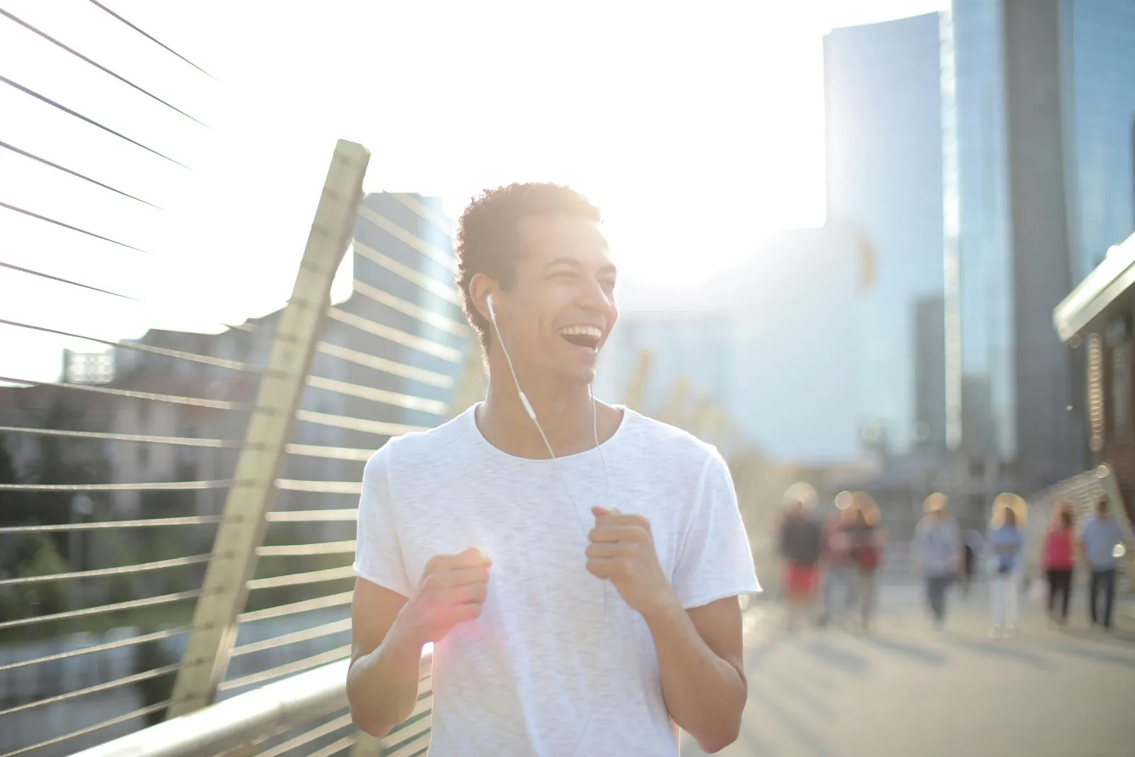 Ein lächelnder junger Mann joggt mit Kopfhörern über eine sonnige Stadtbrücke