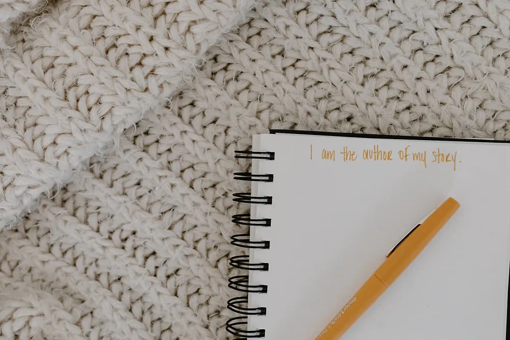 Open spiral notebook on a cream knit blanket with a gold pen and the handwritten line 'I am the author of my story'