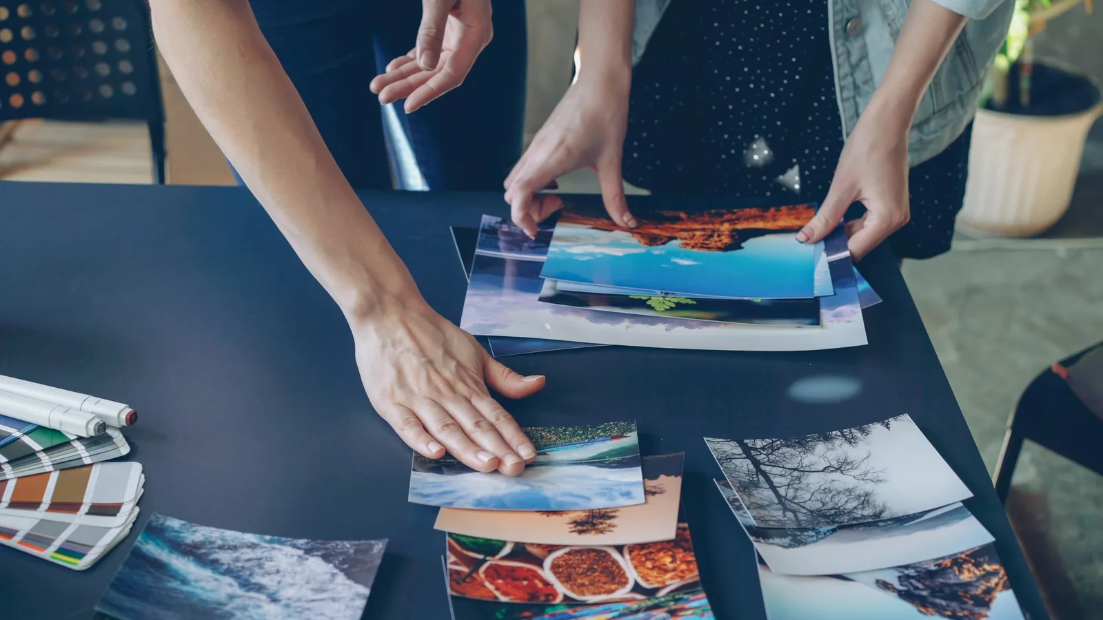 Two people's hands laying printed photos out on a dark table, with color swatches and markers at the edge