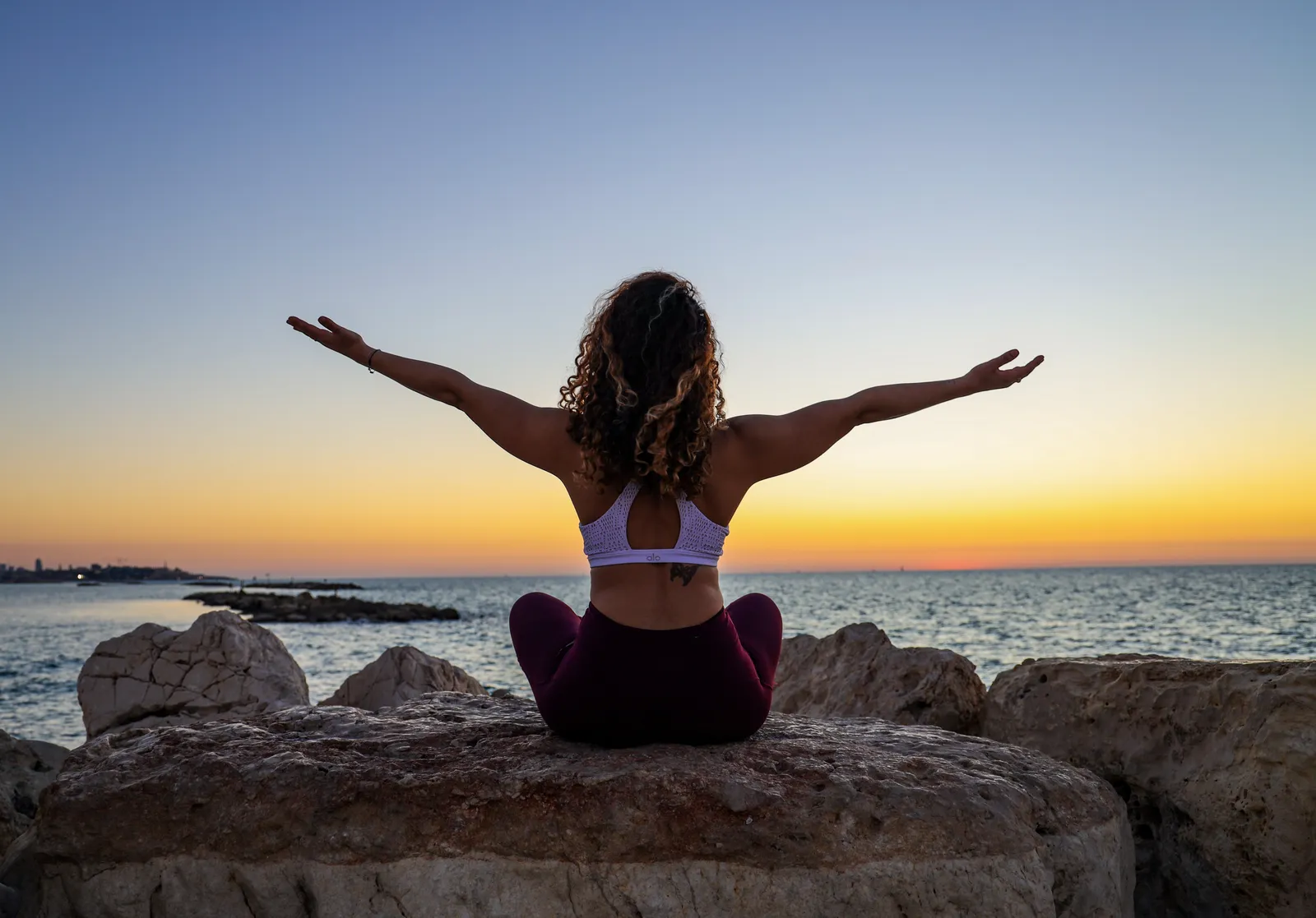 A woman in athletic wear sitting on a rock by the sea at sunset, arms raised in celebration