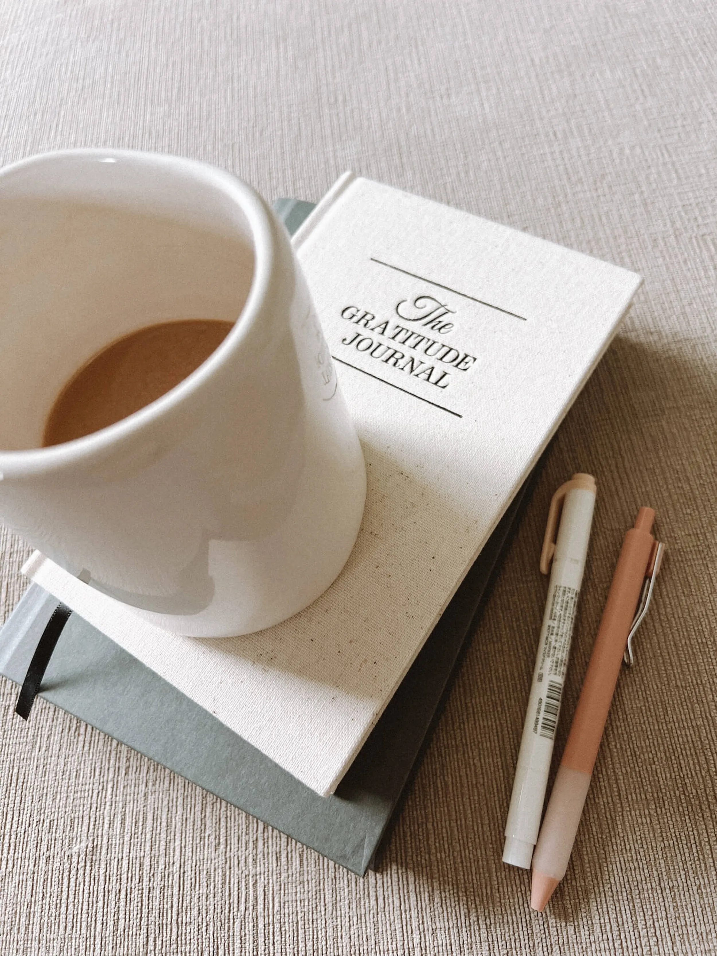 Cremefarbene Tasse Kaffee auf einem Buch mit dem Titel 'The Gratitude Journal' neben zwei Stiften