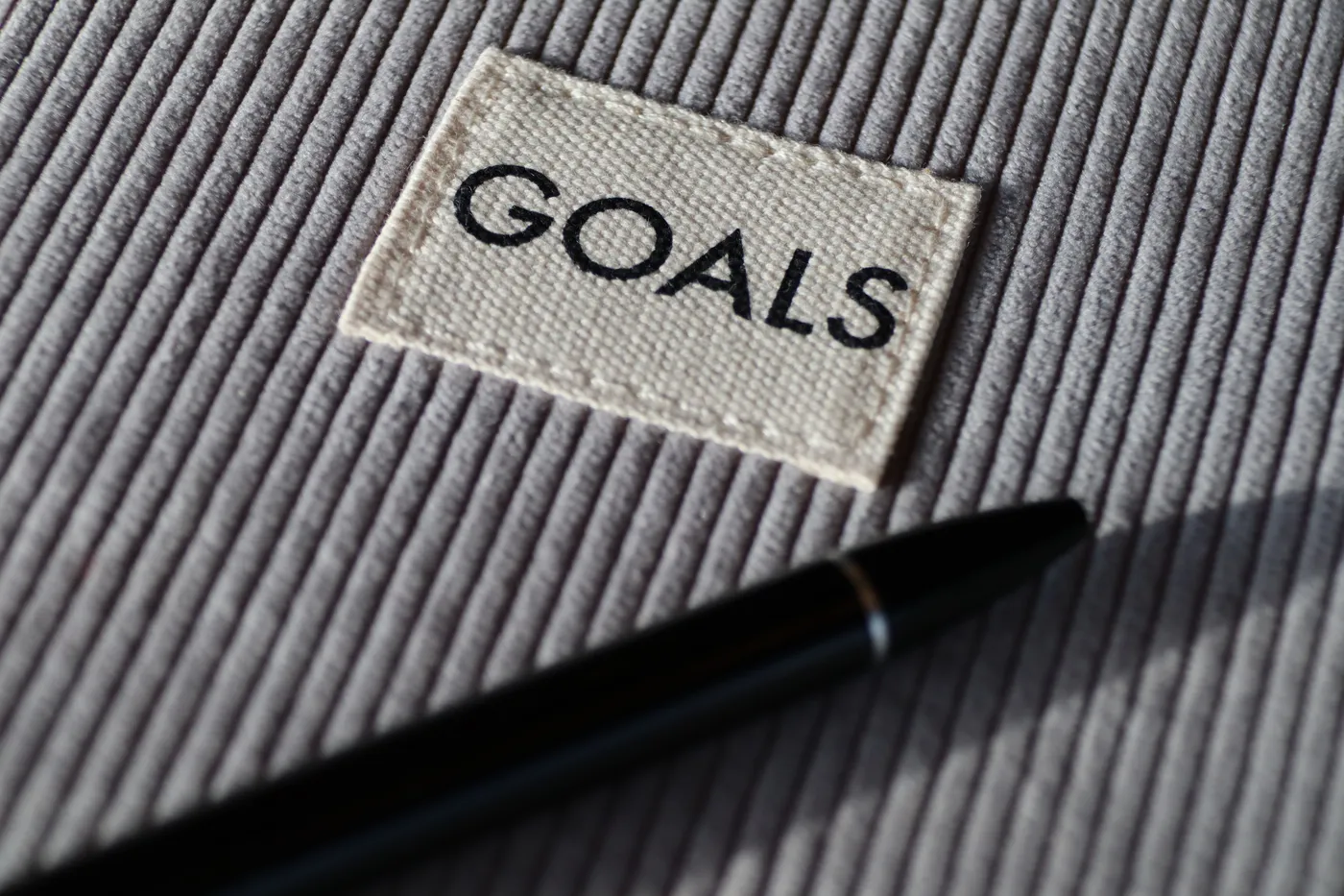Ein Stoffetikett mit der Aufschrift 'GOALS' auf grauem Cordstoff, daneben ein schlanker schwarzer Stift