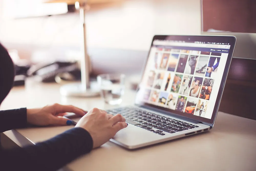 Hands typing on a laptop that shows a grid of inspiration images at a bright, sunlit desk