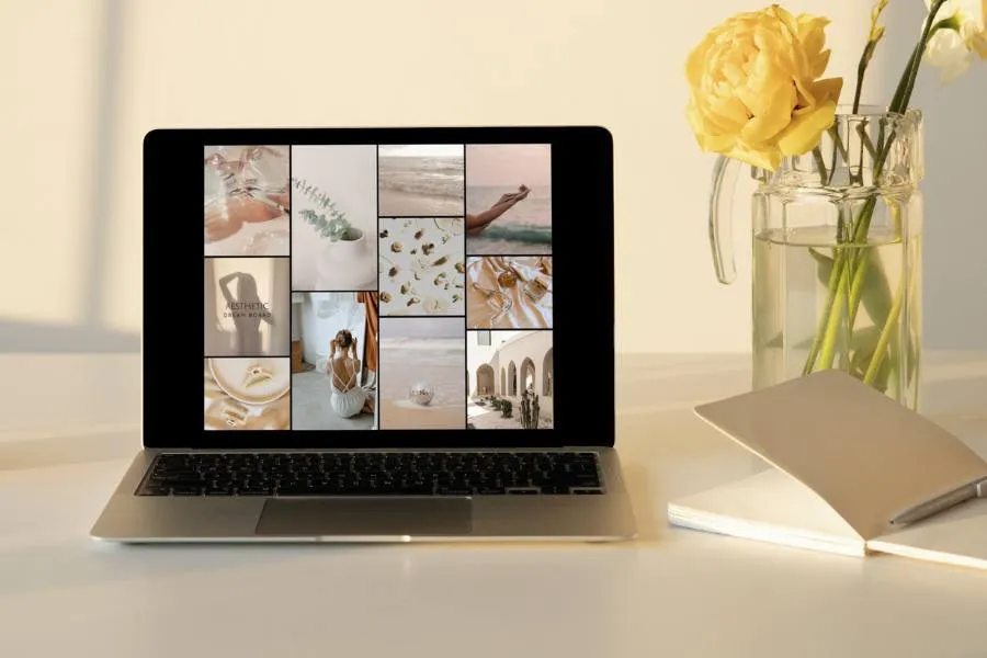 Laptop on a desk displaying an aesthetic digital dream board collage, beside yellow roses and a notebook
