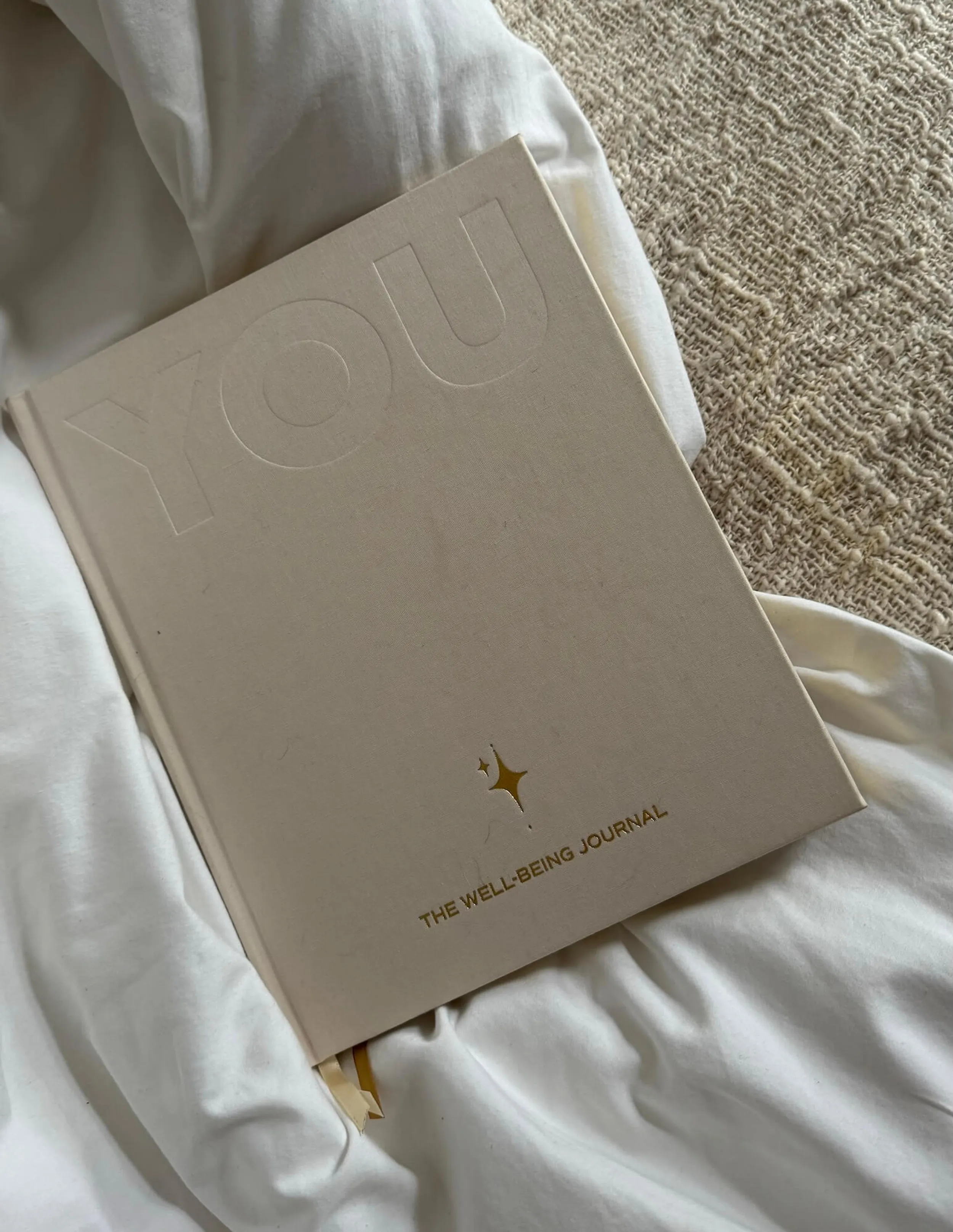 Cream linen journal embossed 'YOU' with gold lettering 'The Well-Being Journal' resting on soft white bedding