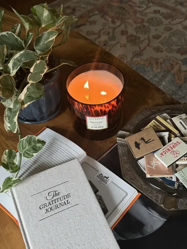 Leinen-Notizbuch 'The Gratitude Journal' neben einer brennenden Kerze und einer Rankpflanze auf dunklem Holztisch