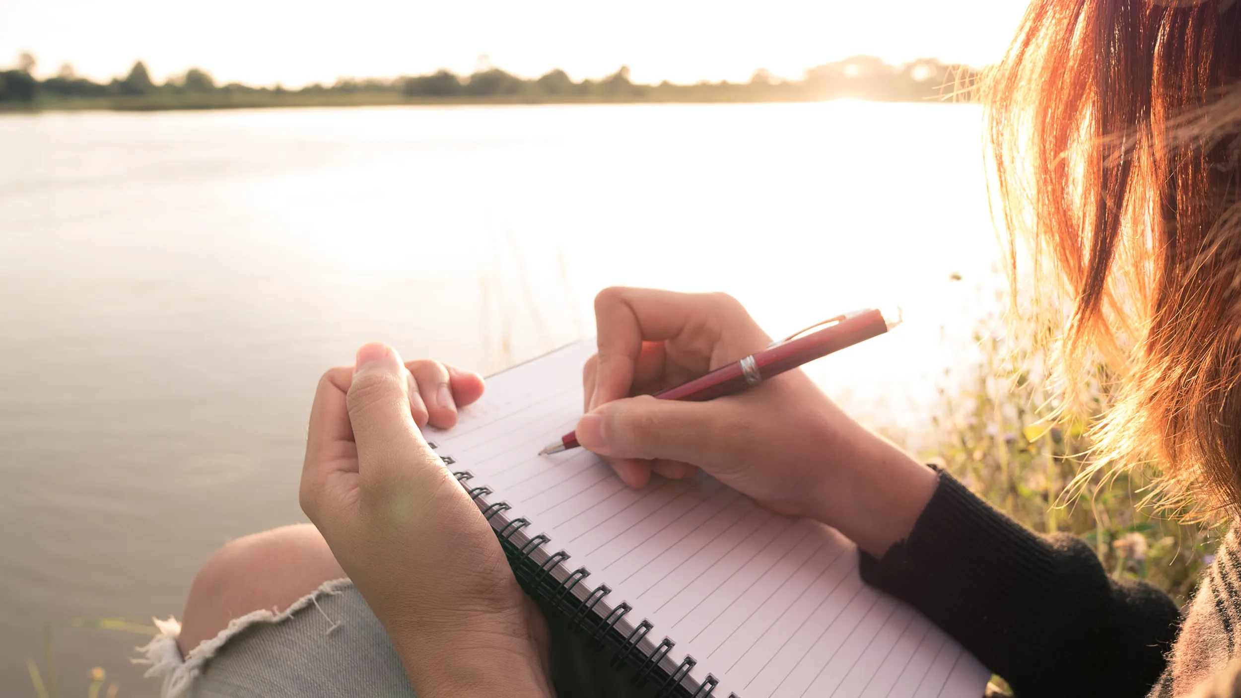 A young woman writing in a journal by the river at dusk, using Dr. Ira Progoff's method