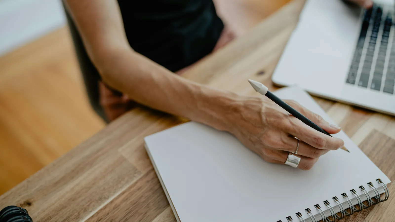 Eine Hand mit Bleistift über einem leeren Notizbuch, daneben ein Laptop auf dem Holztisch