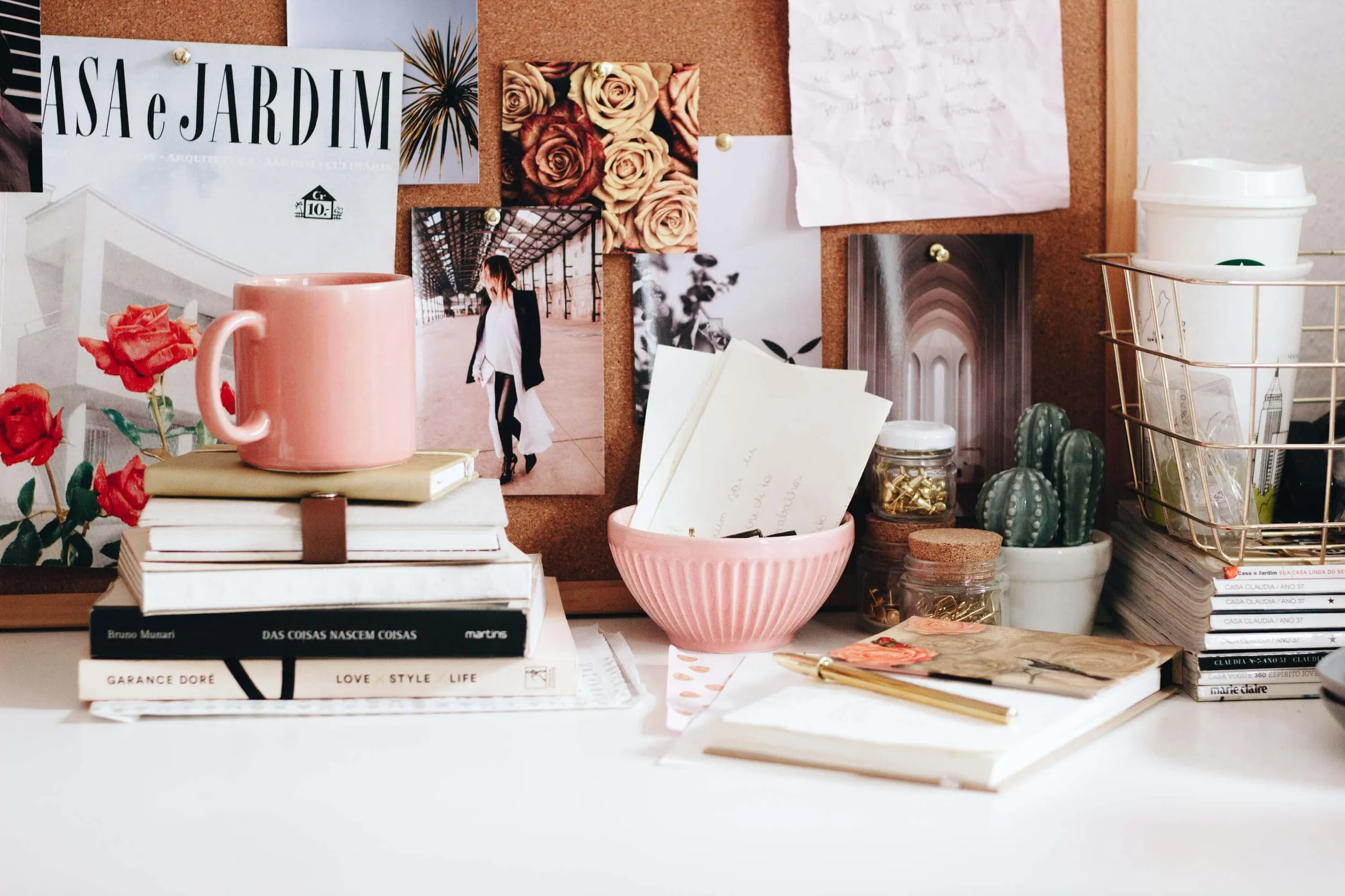 Styled desk with a cork mood board, pink mug on stacked design books and fresh roses