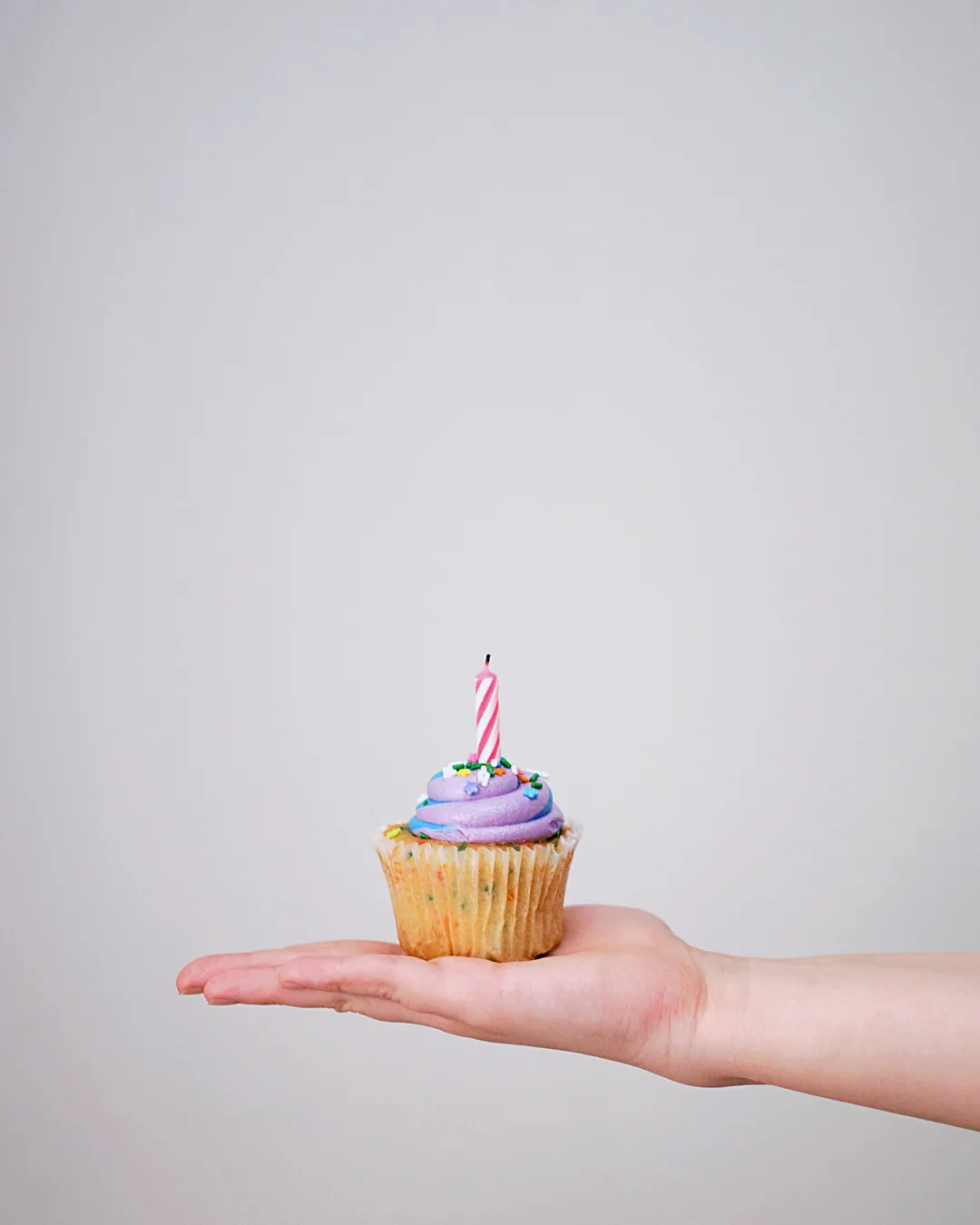 Eine Hand hält einen Cupcake mit einer einzelnen brennenden Kerze vor einem hellgrauen Hintergrund
