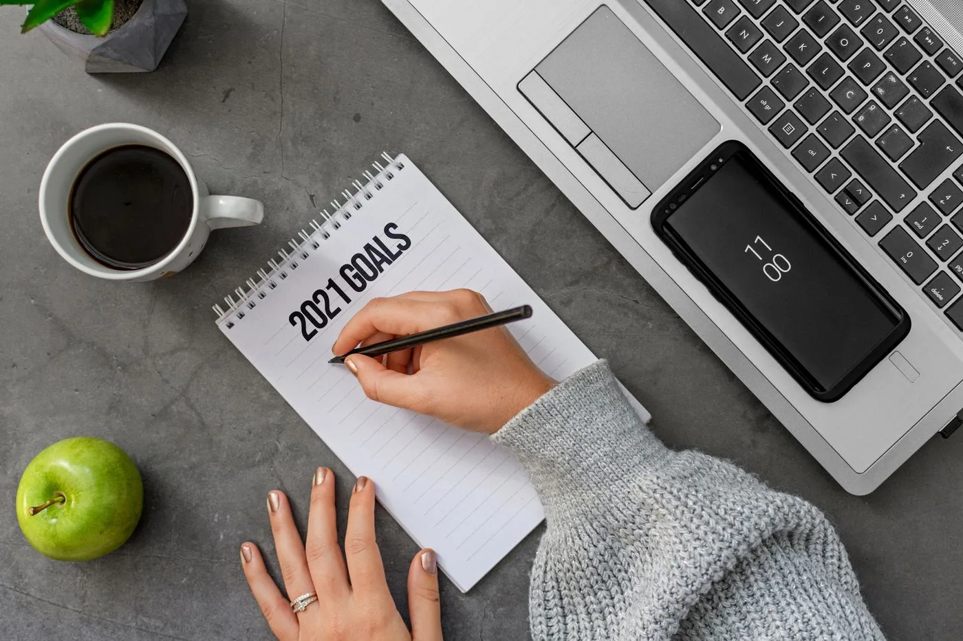A woman writing her goals for the year in a notebook at a desk with a laptop and a cup of coffee