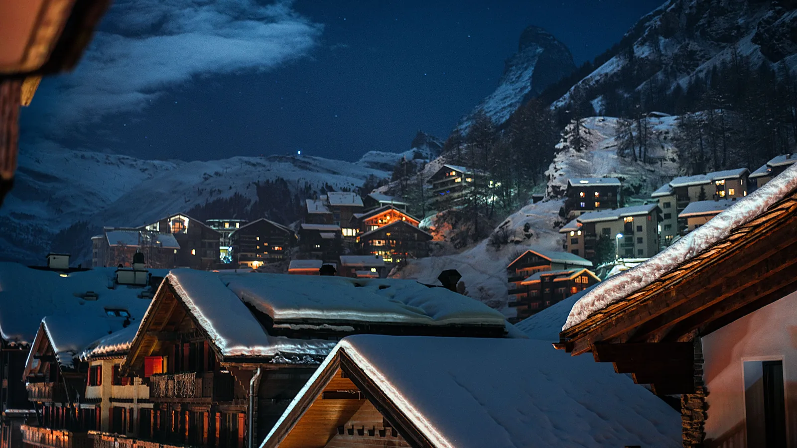 Verschneite Holzhäuser in einem Alpendorf bei Nacht, dahinter die Berge