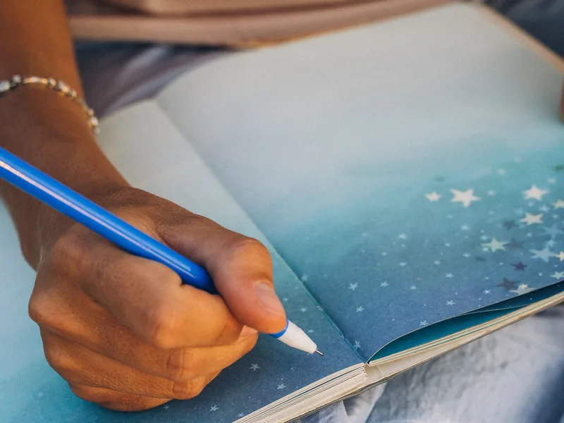 Eine Hand schreibt mit einem blauen Stift in ein Notizbuch mit Sternenhimmel auf den Seiten