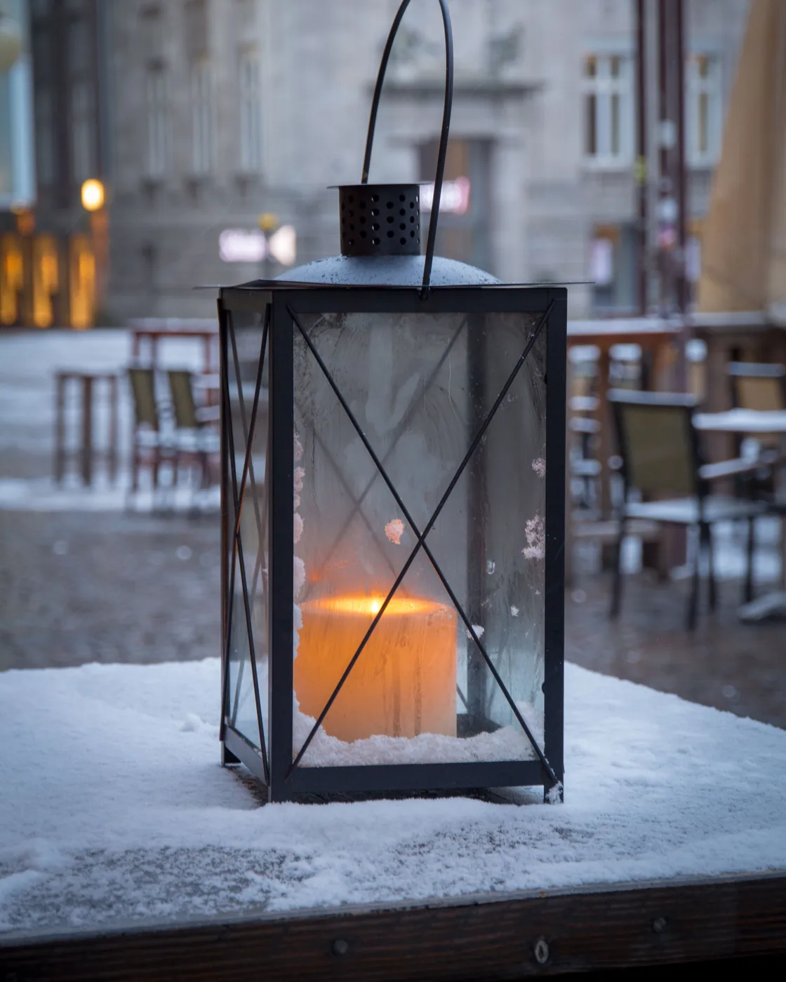Eine Kerze brennt in einer Laterne auf einem verschneiten Tisch vor einem stillen Café