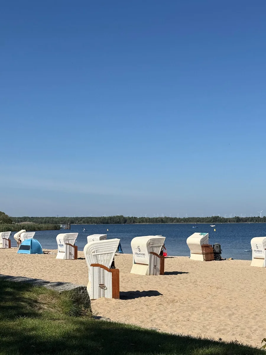 A sandy lake beach with white hooded wicker beach chairs on a summer day