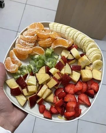 A hand holding a plate of tangerines, kiwi, apples, strawberries and bananas